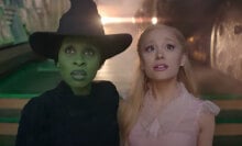 A woman with green skin in a witch's hat stands next to a woman in a pink dress.