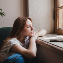 Upset redhead teen girl sitting by window looking at phone waiting call from boyfriend, feeling sad and depressed teenager looking at smartphone wait for message