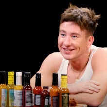 A man in a white vest sits at a table covered in sauce bottles in a dark room.