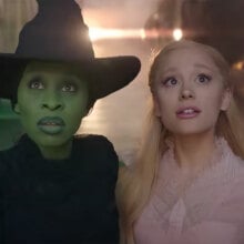 A woman with green skin in a witch's hat stands next to a woman in a pink dress.