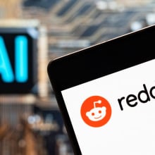 A phone displaying the Reddit logo in front of a screen that reads "AI". 