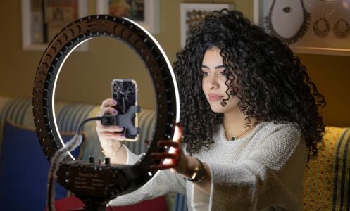 A woman with big, curly hair sits in front of her iPhone and a large ring light.