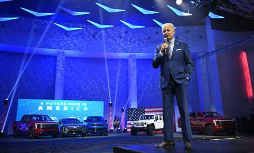 Old white man in blue suit speaks into a microphone. In the background is a display lined with electric vehicles in red, white, and blue variations