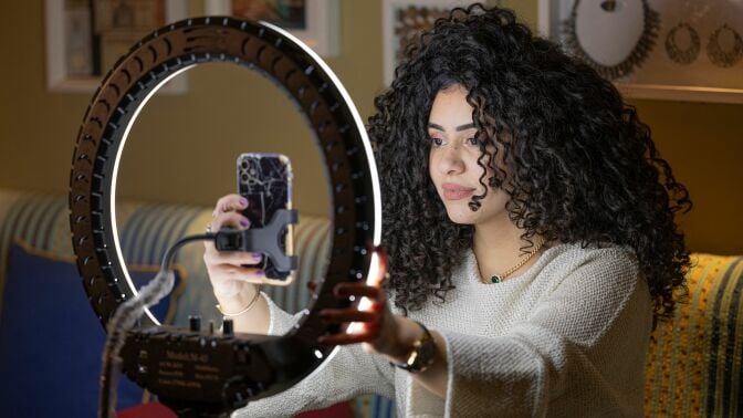 A woman with big, curly hair sits in front of her iPhone and a large ring light.