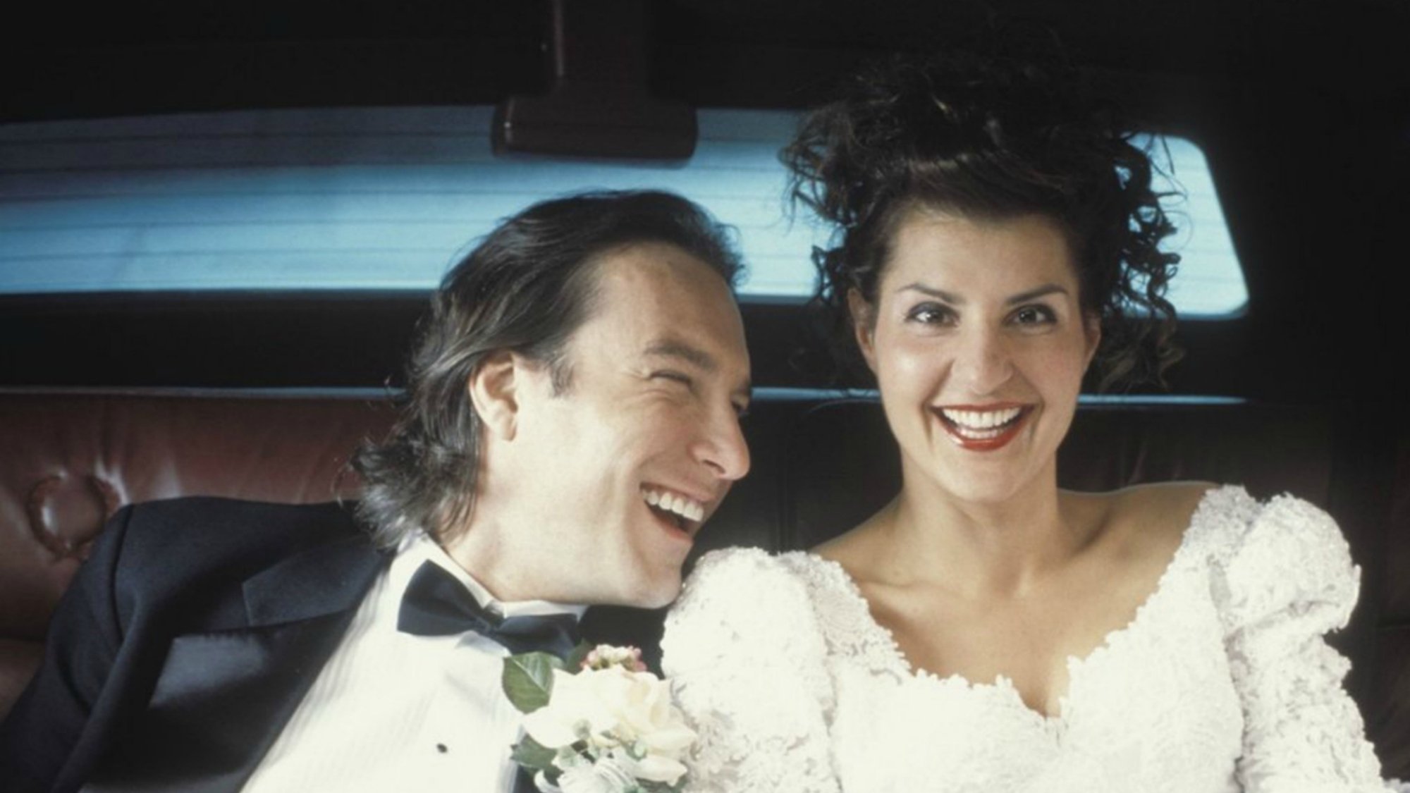 A woman in a wedding gown and a man in a tuxedo sit in the back seat of a car.