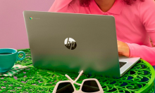 a woman working on a HP Chromebook 14a-ne0013dx at a green table that has a cup of tea and a pair of sunglasses sitting on it