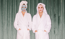 two people in robes and skincare face masks looking at the camera